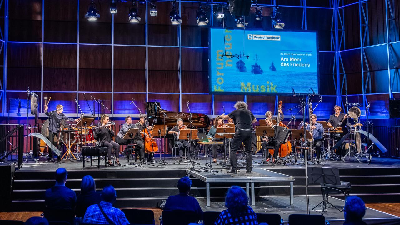 Ein Orchester steht auf der bläulich illuminierten Bühne des Kammermusiksaales im Deutschlandfunk Köln mit Publikum.