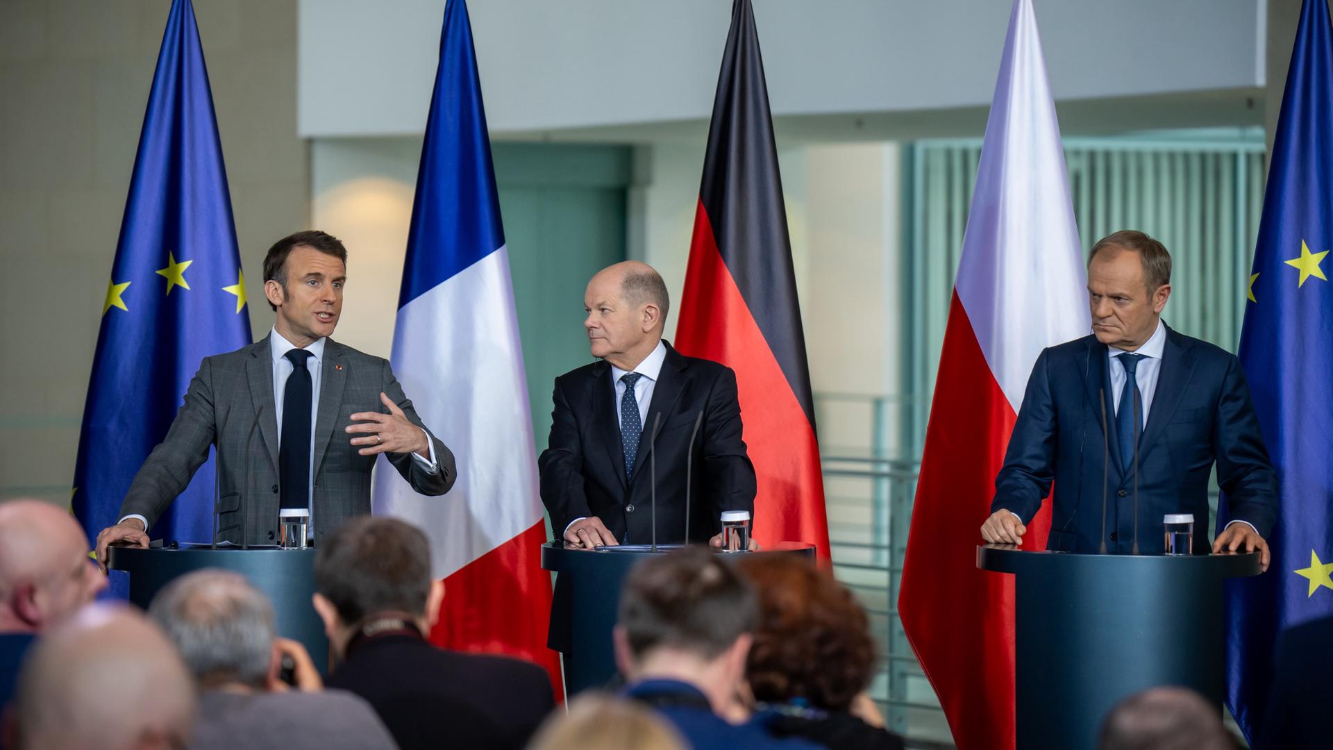 Bundeskanzler Scholz, Frankreichs Präsident Macron und Polens Ministerpräsident Tusk bei einer gemeinsamen Pressekonferenz. Im Hintergrund die jeweiligen Nationalflaggen und die Europa-Flagge. 