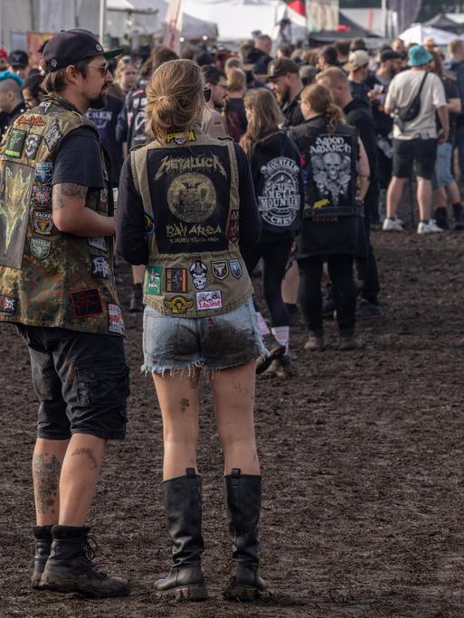 Mehrere Menschen stehen beim Wacken-Festival auf matschigem Boden. Sie tragen Heavy Metal-Kleidung.
