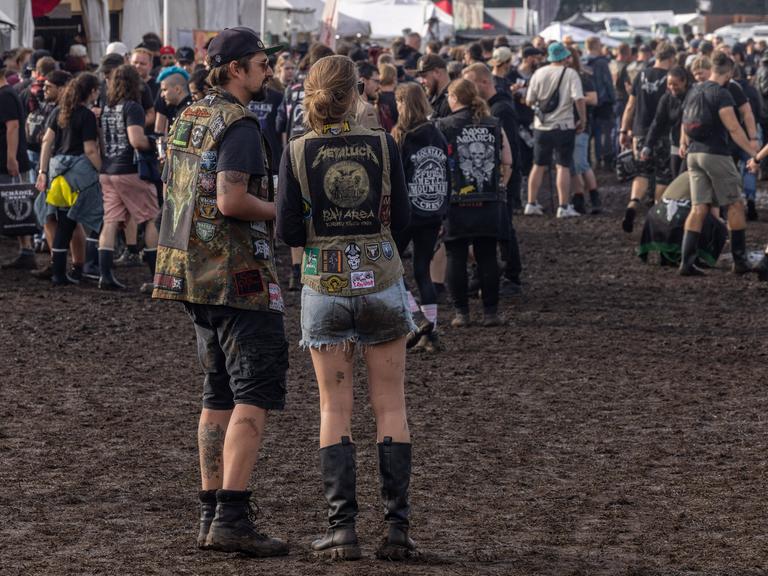 Mehrere Menschen stehen beim Wacken-Festival auf matschigem Boden. Sie tragen Heavy Metal-Kleidung.