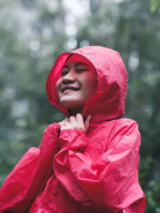 Eine junge Frau spaziert in einer roten Regenjacke durch einen Wald. Es regnet und sie lächelt.