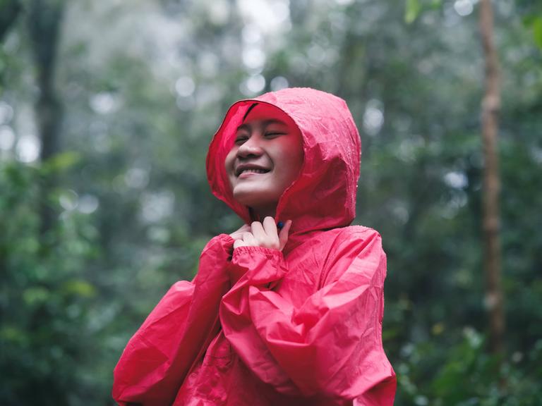 Eine junge Frau spaziert in einer roten Regenjacke durch einen Wald. Es regnet und sie lächelt.