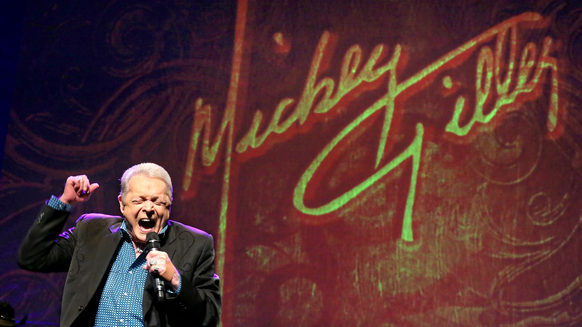 Ein weißer alter Mann singt mit weitgeöffnetem Mund und rechter erhobener Faus auf einer Bühne. Im Hintergrund steht an der Wand in Schreibschrift sein Name Mickey Gilley Ein weißer alter Mann singt mit weitgeöffnetem Mund und rechter erhobener Faus auf einer Bühne. Im Hintergrund steht an der Wand in Schreibschrift sein Name Mickey Gilley