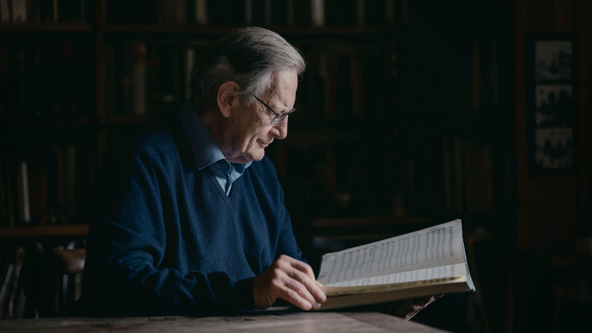 Sir John Eliot Gardiner sitzt an einem Schreibtisch und blickt in eine große, aufgeschlagene Partitur. 