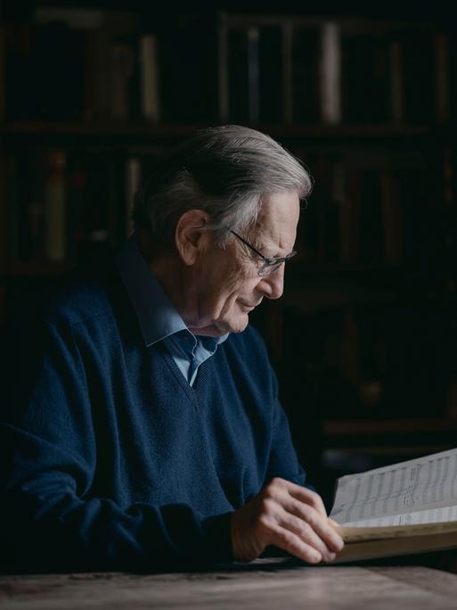 Sir John Eliot Gardiner sitzt an einem Schreibtisch und blickt in eine große, aufgeschlagene Partitur. 