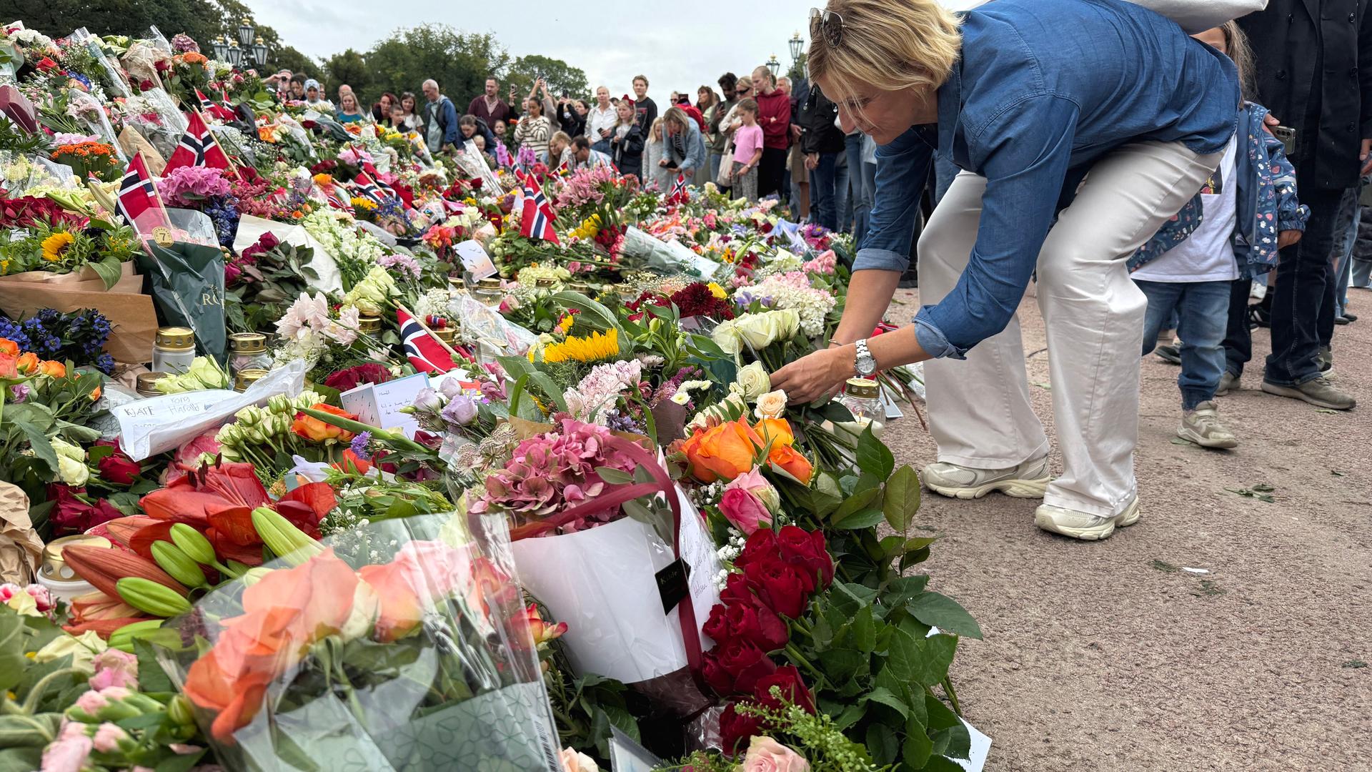 Eine riesige Menge Blumen liegt an einem Zaun. Eine Frau legt weitere Blumen dazu.
