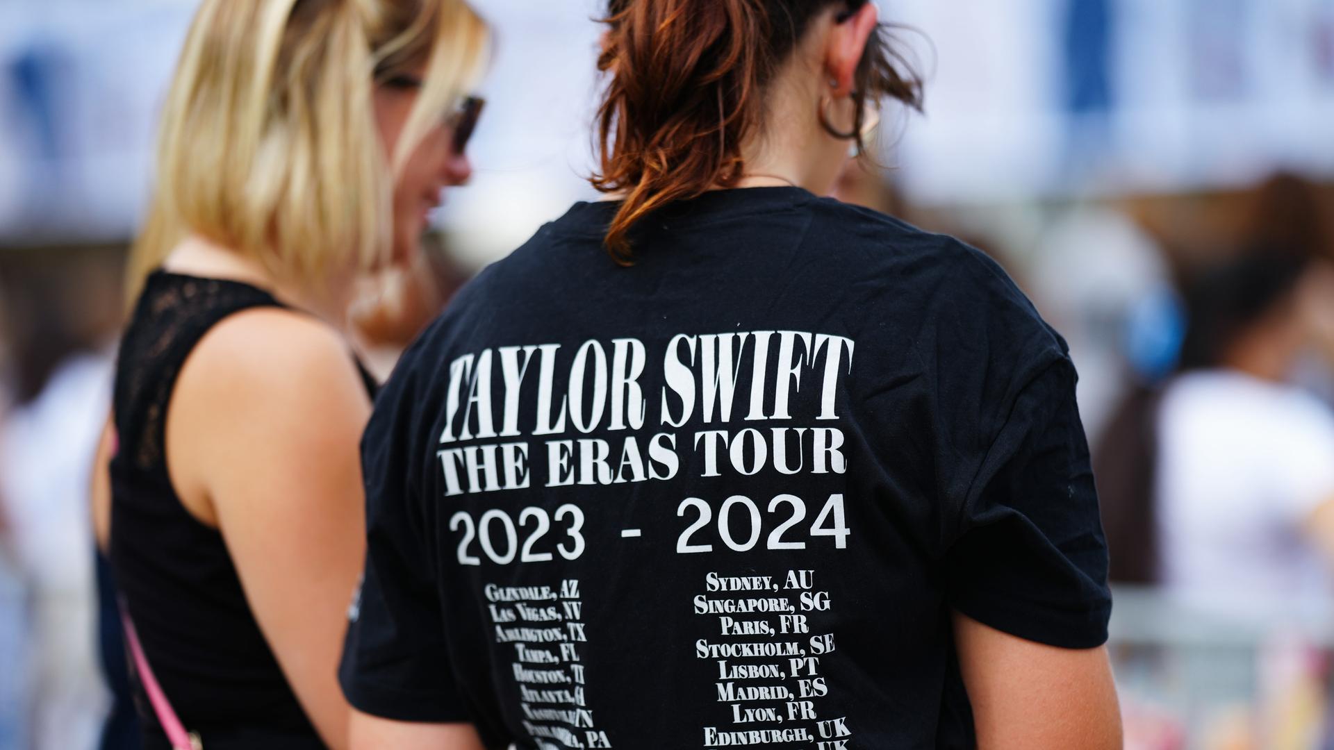 Zwei Frauen sind von hinten zu sehen. Die rechte hat ein schwarzes Shirt mit den Tourdaten von Taylor Swift an. 