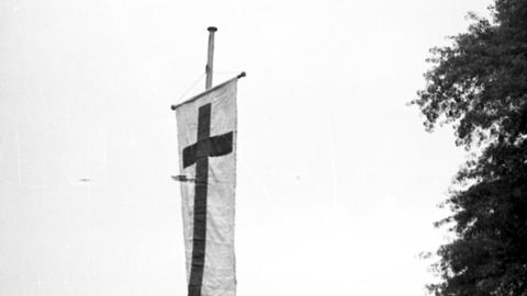 Ein Mann hisst eine weiße Fahne mit Kreuz auf einem Feld während des evangelischen Kirchentages in Leipzig. (Symbolbild)