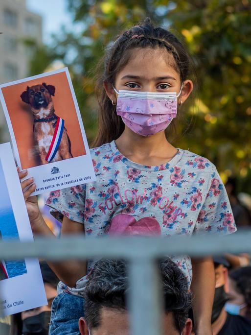 Ein kleines Mädchen mit rosa Mundschutz sitzt auf der Schulter ihres Vaters und beide halten jeweils ein Foto von einem Hund bzw. dem neuen Präsidenten Boric mit einer gestreiften Schärpe hoch.