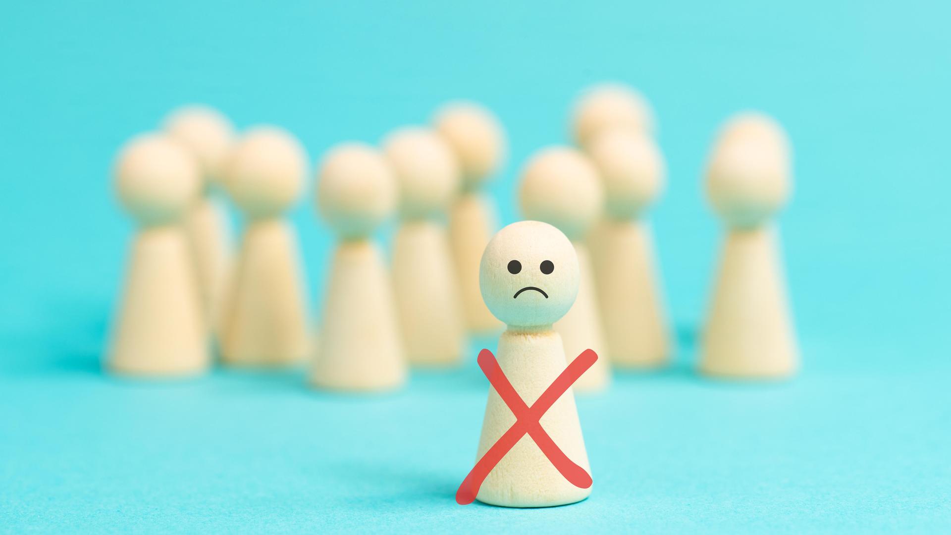 Einsame Holzfigur mit traurigem Smiley-Gesicht steht vor einer anonymen Menge gesichtsloser Holzfiguren als Symbolbild für Ausgrenzung, Mobbing und Demütigung. Einsame Holzfigur mit traurigem Smiley-Gesicht steht vor einer anonymen Menge gesichtsloser Holzfiguren als Symbolbild für Ausgrenzung, Mobbing und Demütigung.
