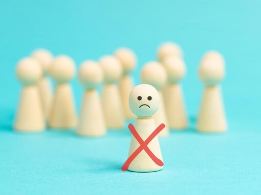 Einsame Holzfigur mit traurigem Smiley-Gesicht steht vor einer anonymen Menge gesichtsloser Holzfiguren als Symbolbild für Ausgrenzung, Mobbing und Demütigung.