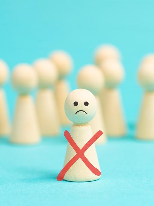 Einsame Holzfigur mit traurigem Smiley-Gesicht steht vor einer anonymen Menge gesichtsloser Holzfiguren als Symbolbild für Ausgrenzung, Mobbing und Demütigung. Einsame Holzfigur mit traurigem Smiley-Gesicht steht vor einer anonymen Menge gesichtsloser Holzfiguren als Symbolbild für Ausgrenzung, Mobbing und Demütigung.