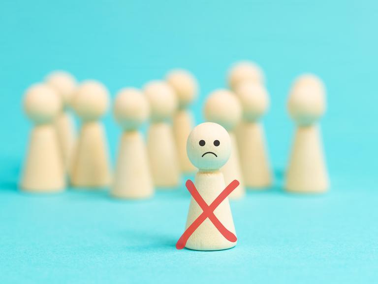 Einsame Holzfigur mit traurigem Smiley-Gesicht steht vor einer anonymen Menge gesichtsloser Holzfiguren als Symbolbild für Ausgrenzung, Mobbing und Demütigung. Einsame Holzfigur mit traurigem Smiley-Gesicht steht vor einer anonymen Menge gesichtsloser Holzfiguren als Symbolbild für Ausgrenzung, Mobbing und Demütigung.