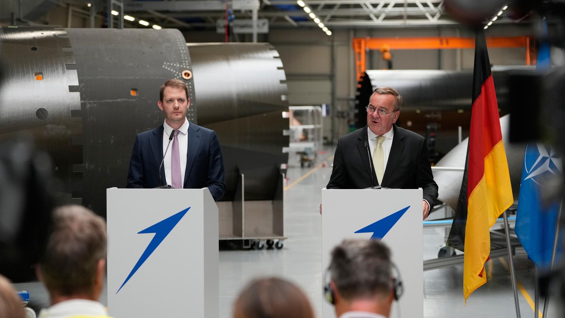 Verteidigungsminister Pistorius (r.) und Isar-Aerospace-Chef Metzler stehen in einer Halle an Rednerpulten - Pistorius spricht.