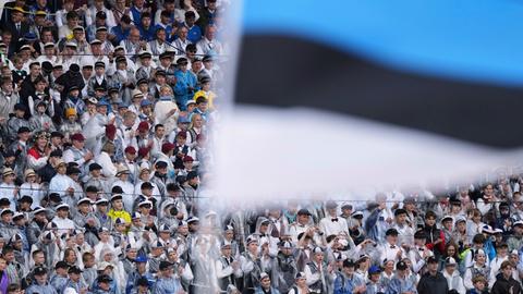 Chorsänger sitzen beim jährlichen Liederfestival in Tallinn in einem Stadion. Vor ihnen im Bildvordergrund wird eine estnische Flagge geschwenkt.