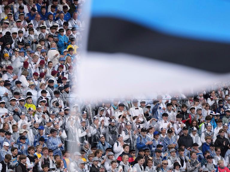 Chorsänger sitzen beim jährlichen Liederfestival in Tallinn in einem Stadion. Vor ihnen im Bildvordergrund wird eine estnische Flagge geschwenkt.