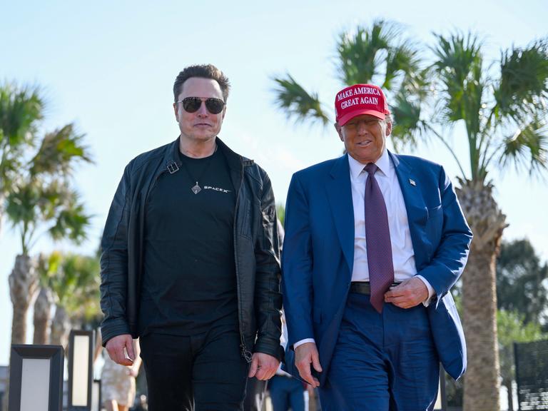 US-Präsident Donald Trump auf der rechten und sein Vertrauter Elon Musk auf der linken Seite laufen vor Palmen auf die Kamera zu. Trump trägt ein MAGA-Basecap.