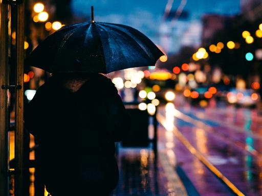 Abendstimmung in einer verregneten Großstadt. Im Vordergrund steht ein Mensch mit Regenschirm.