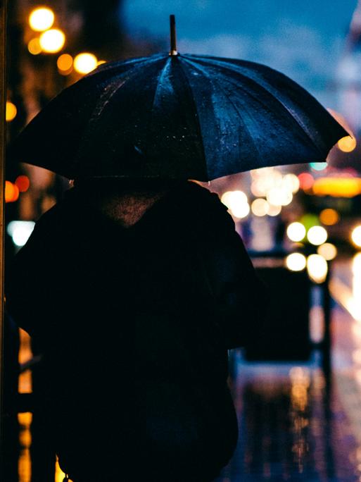 Abendstimmung in einer verregneten Großstadt. Im Vordergrund steht ein Mensch mit Regenschirm.