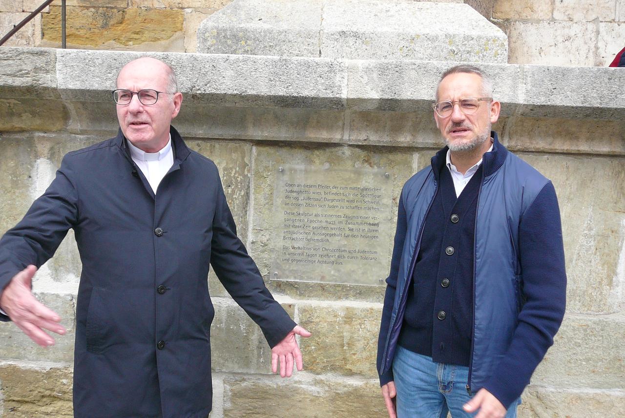 Zwei Männer mittleren Alters stehen vor dem Schild, das am Regensburger Dom den historischen Kontext zum "Judensau"-Relief liefert.