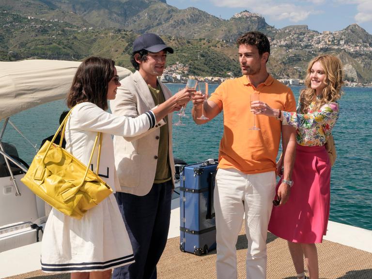 Szene aus der zweiten Staffel der Serie "The White Lotus". Es stehen zwei gut gekleidete Paare auf einem Bootsanleger in Italien und stoßen mit Sektgläsern an.