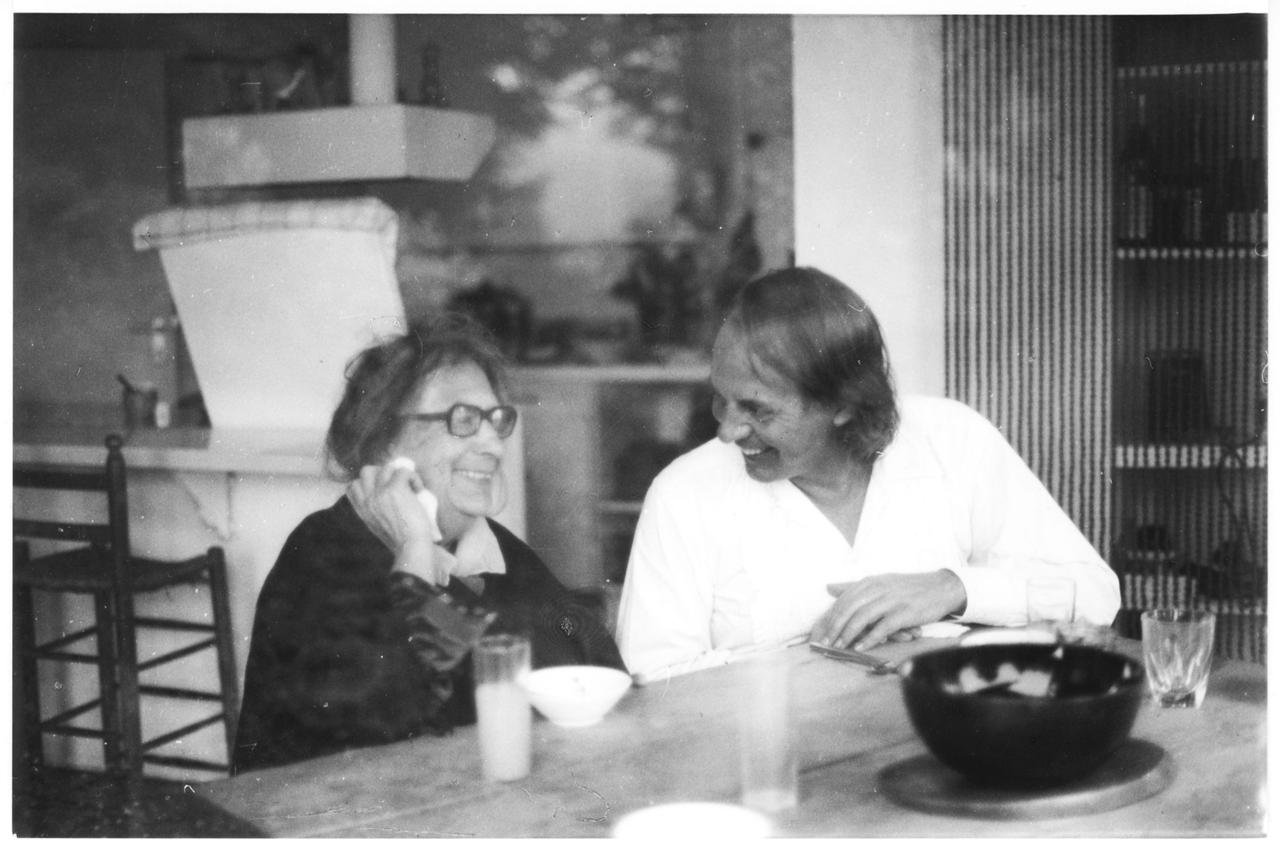 Der weiß gekleidete, jüngere Karlheinz Stockhausen sitzt neben der älteren Recha Freier, beide lachend ins Gespräch vertieft.