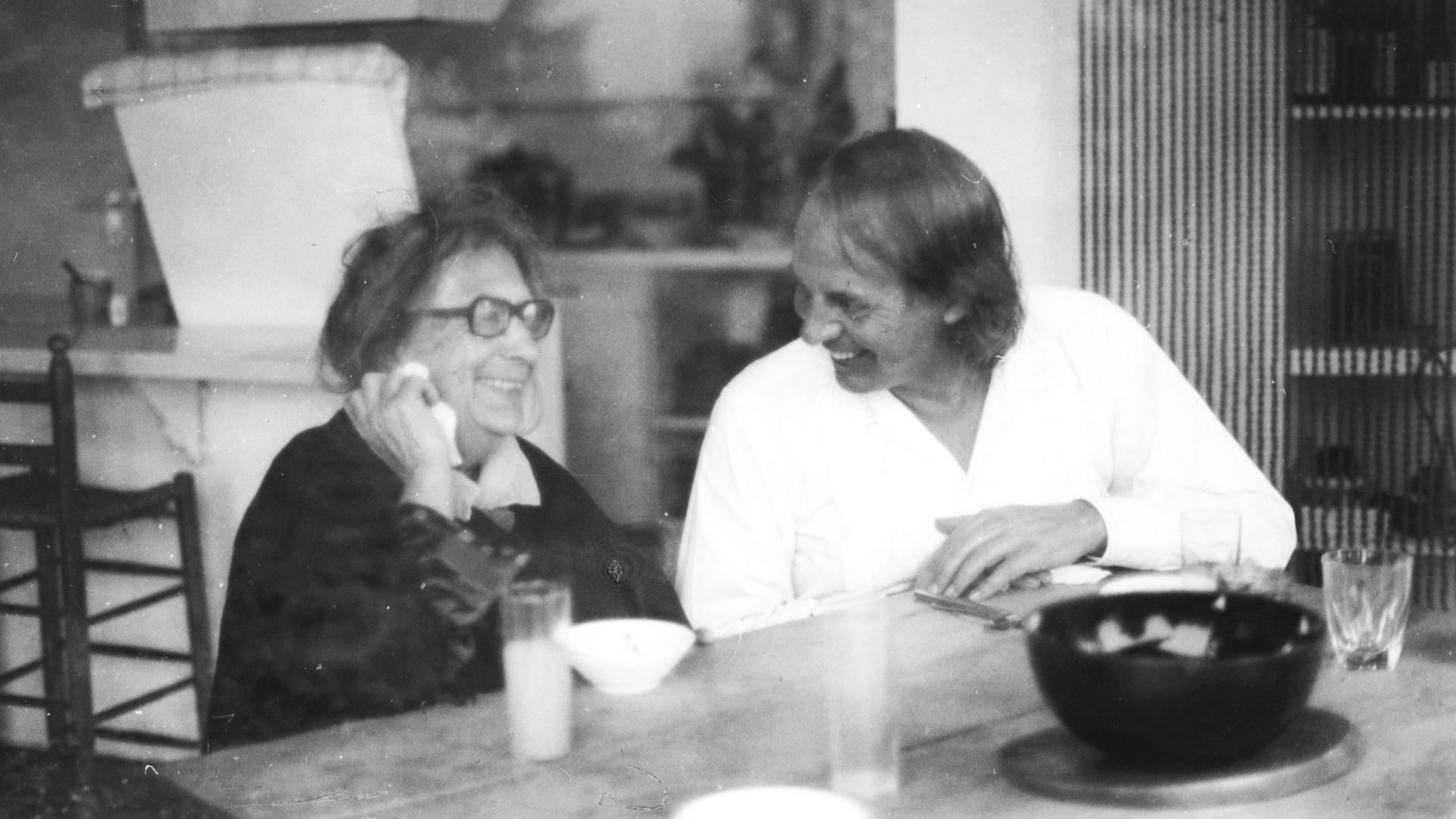 Der weiß gekleidete, jüngere Karlheinz Stockhausen sitzt neben der älteren Recha Freier, beide lachend ins Gespräch vertieft.