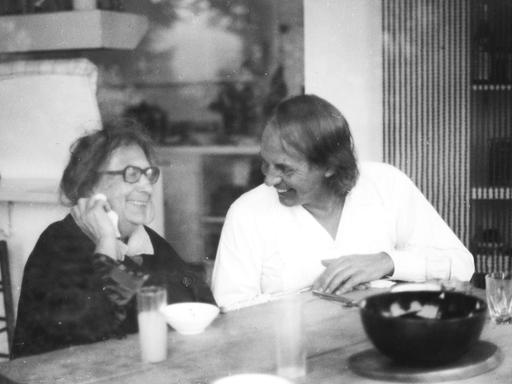 Der weiß gekleidete, jüngere Karlheinz Stockhausen sitzt neben der älteren Recha Freier, beide lachend ins Gespräch vertieft.