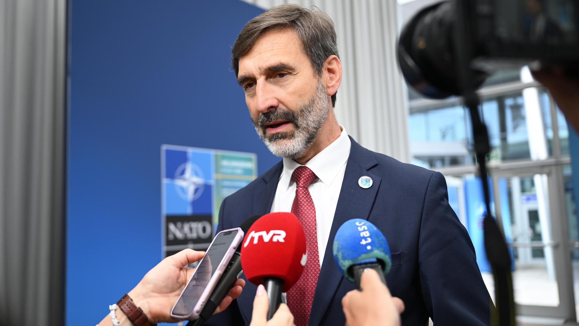 Der Außenminister der Slowakei, Juraj Blanar, spricht in zwei Mikrofone und ein Handy von Reportern. Dahinter an einer Wand ein NATO-Emblem. 