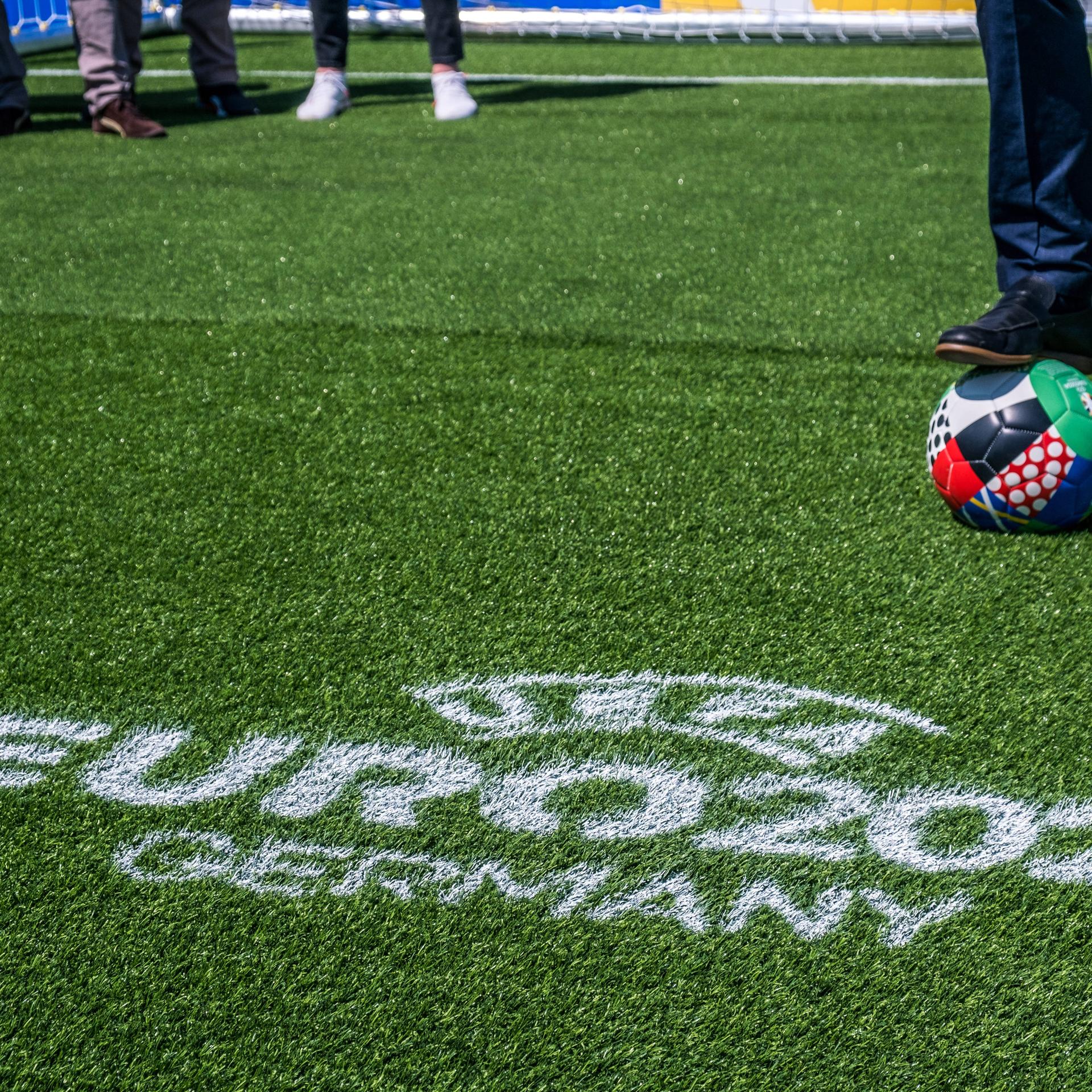 Das Bild zeigt einen Fußball-Rasen, auf dem mit weißer Farbe "UEFA EURO 2024 Germany" geschrieben ist, im Hintergrund ist der Ball für die Europameisterschaft in Deutschland zu sehen.