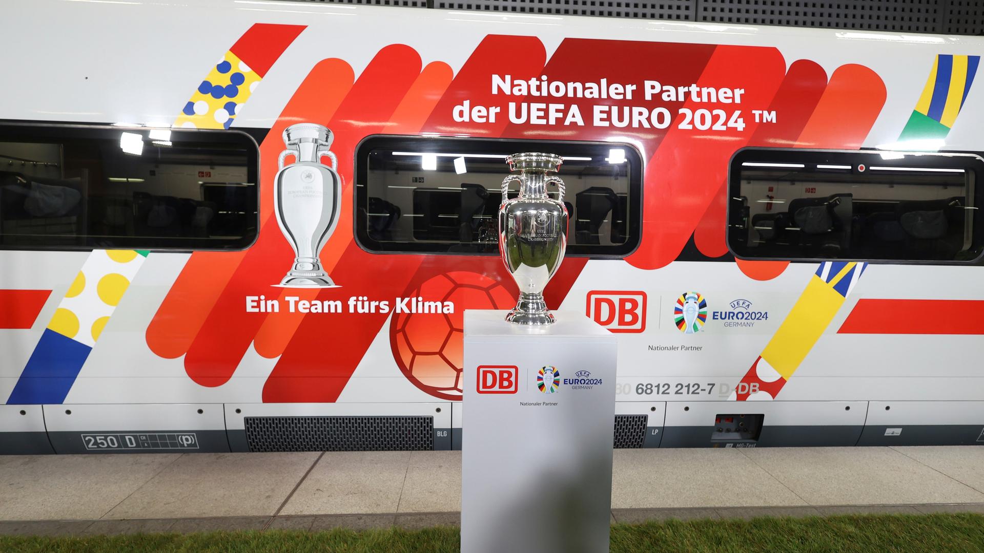Ein Zug der Deutschen Bahn ist mit dem Schriftzug "Nationaler Partner der UEFA Euro 2024" beklebt, davor steht der Pokal auf einem Podest. Ein Zug der Deutschen Bahn ist mit dem Schriftzug "Nationaler Partner der UEFA Euro 2024" beklebt, davor steht der Pokal auf einem Podest.