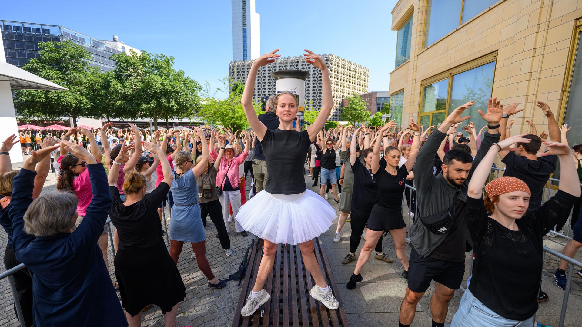 Eine Person in Ballettpose steht auf einer Sitzbank und um sie herum sind zahlreichen Menschen auf der Straße ebenfalls in Ballettpose