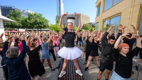 Eine Person in Ballettpose steht auf einer Sitzbank und um sie herum sind zahlreichen Menschen auf der Straße ebenfalls in Ballettpose