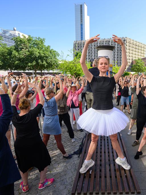 Eine Person in Ballettpose steht auf einer Sitzbank und um sie herum sind zahlreichen Menschen auf der Straße ebenfalls in Ballettpose