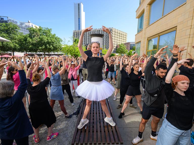Eine Person in Ballettpose steht auf einer Sitzbank und um sie herum sind zahlreichen Menschen auf der Straße ebenfalls in Ballettpose