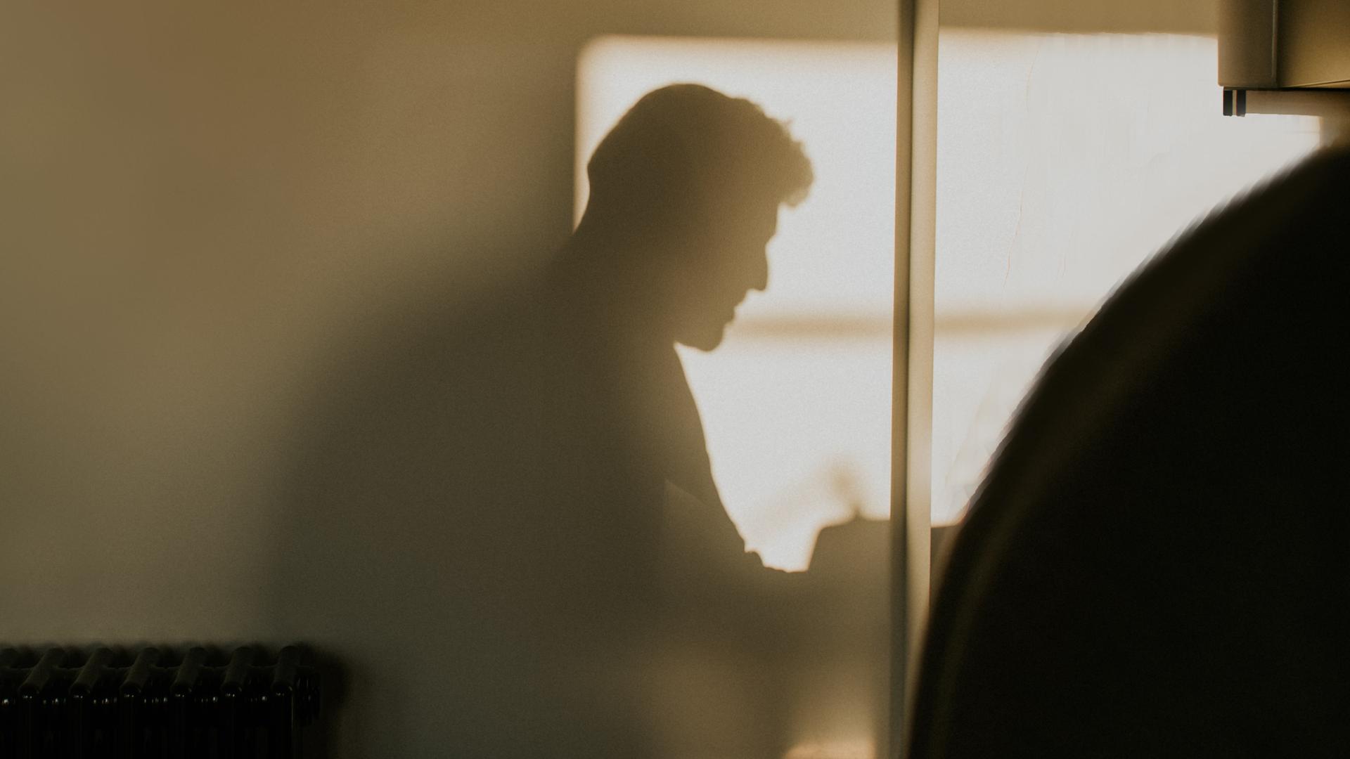 Der Schatten einer männlich gelesenen Person bildet sich an einer Wand ab. Der Schatten einer männlich gelesenen Person bildet sich an einer Wand ab.