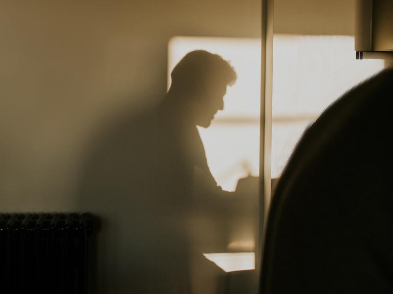 Der Schatten einer männlich gelesenen Person bildet sich an einer Wand ab. Der Schatten einer männlich gelesenen Person bildet sich an einer Wand ab.