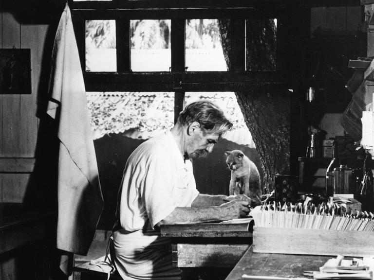 Historisches Schwarzweißfoto von Albert Schweitzer, der in einem weißen Hemd am Schreibtisch in seinem Büro sitzt. Neben ihm auf dem Tisch sitzt eine Katze.