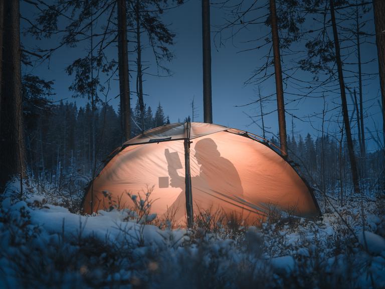 Eine Person sitzt in einer Winterlandschaft in einem Zelt und liest ein Buch bei Taschenlampenschein. Der Schatten ist von außen zu sehen.