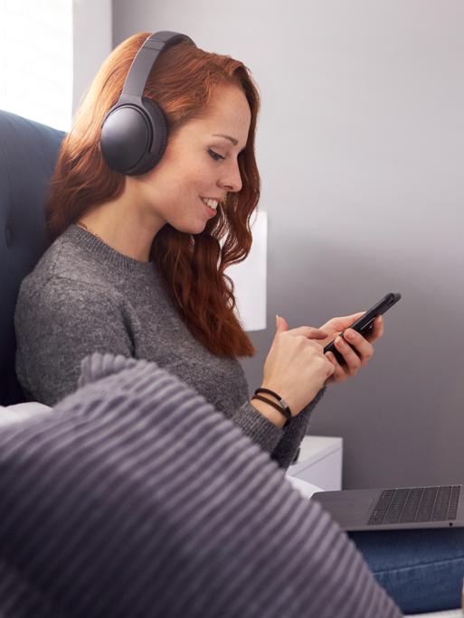 Ein junge, rothaarige Frau sitzt auf dem Sofa und schaut auf ein Smartphone. Sie trägt Kopfhörer und lächelt, offenbar wählt sie einen Audio-Titel aus. Auf ihrem Schoß steht ein geöffneter Laptop. 