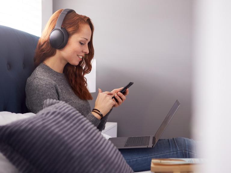Ein junge, rothaarige Frau sitzt auf dem Sofa und schaut auf ein Smartphone. Sie trägt Kopfhörer und lächelt, offenbar wählt sie einen Audio-Titel aus. Auf ihrem Schoß steht ein geöffneter Laptop. 