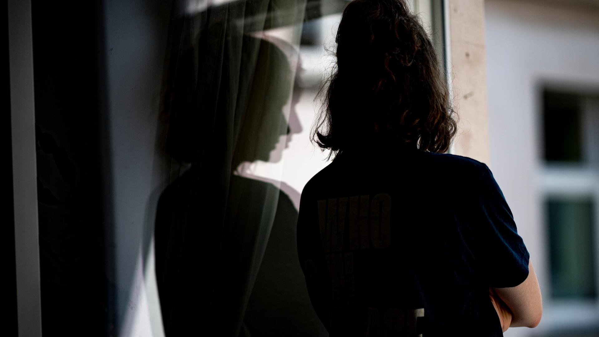 Eine Frau steht mit verschränkten Armen in ihrer Wohnung an einem Fenster.