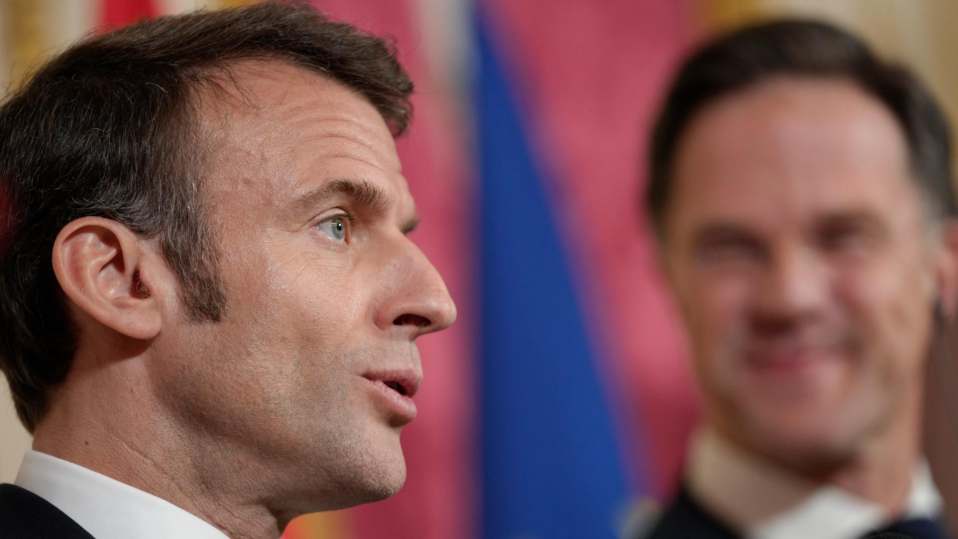 Macrons Kopf ist groß in der linken Bildhälfte von der Seite zu sehen; er sagte etwas. In der rechten Bildhälfte unscharf von vorne Ruttes Kopf; der Ministerpräsident lächelt. Macrons Kopf ist groß in der linken Bildhälfte von der Seite zu sehen; er sagte etwas. In der rechten Bildhälfte unscharf von vorne Ruttes Kopf; der Ministerpräsident lächelt.