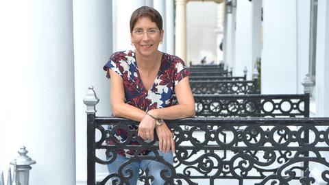 Eine Frau lehnt an einem Geländer. Sie trägt ein T-Shirt mit blau-rot-weißem Blumenmuster, eine randlose Brille, dezenten Goldschmuck und lächelt in die Kamera. Im Hintergrund sind Säulen zu sehen, die an ein herrschaftliches Gebäude erinnern.