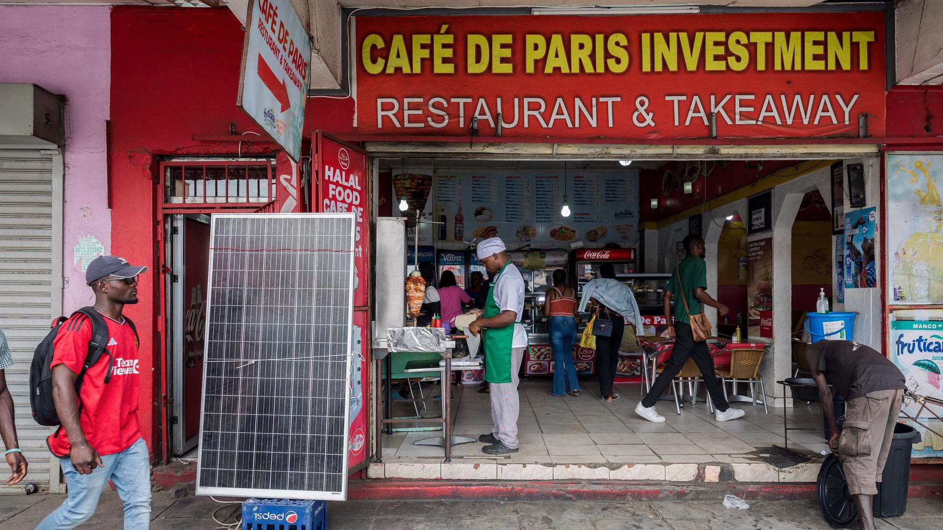 Zu sehen sind Passanten und Mitarbeiter vor einem Geschäft in Sambias Hauptstadt Lusaka. Es trägt den Namen Café De Paris Investment Restaurant & Takeaway.