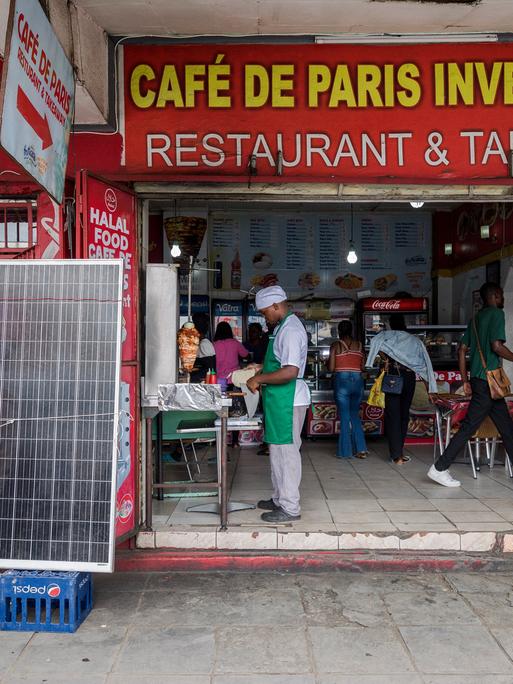 Zu sehen sind Passanten und Mitarbeiter vor einem Geschäft in Sambias Hauptstadt Lusaka. Es trägt den Namen Café De Paris Investment Restaurant & Takeaway.