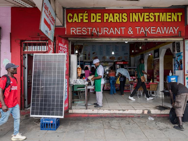 Zu sehen sind Passanten und Mitarbeiter vor einem Geschäft in Sambias Hauptstadt Lusaka. Es trägt den Namen Café De Paris Investment Restaurant & Takeaway.