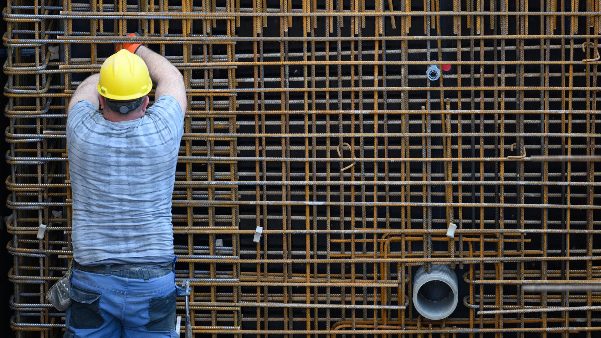 Ein Bauarbeiter mit Schutzkleidung und Helm steht auf einer Baustelle vor einem Metallgitter.