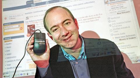 Jeff Bezos im Jahr 2000. Er lächelt in die Kamera und hält eine Computermaus nach oben, auf der Amazon steht. Im Hintergrund ist auf einer Leinwand die Amazon-Webseite zu sehen.