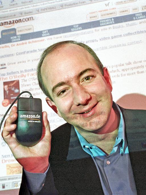Jeff Bezos im Jahr 2000. Er lächelt in die Kamera und hält eine Computermaus nach oben, auf der Amazon steht. Im Hintergrund ist auf einer Leinwand die Amazon-Webseite zu sehen.